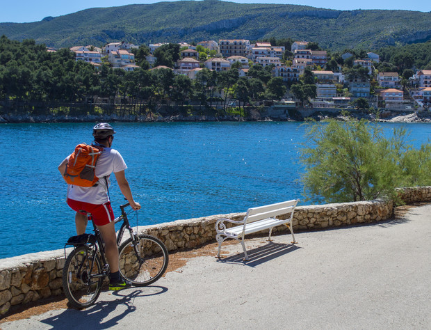 A cyclist pauses, gazing at a picturesque coastal town across the blue water. A white bench and dense trees frame the scene, with distant hills under a clear sky.